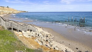 A beach with a wooden walkway leading to a cliff, rocky shoreline, and wooden posts in the water. - Oceans Edge by Wine Coast Holidays (ALDINGA BEACH)