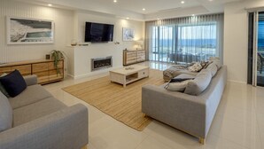 A modern living room with a sofa set, a coffee table, and a flat-screen TV mounted on the wall. - Oceans Edge by Wine Coast Holidays (ALDINGA BEACH)
