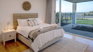 A modern bedroom with a large bed, a nightstand, a decorative wall piece, and a view of a landscaped garden through a glass door. - Oceans Edge by Wine Coast Holidays (ALDINGA BEACH)