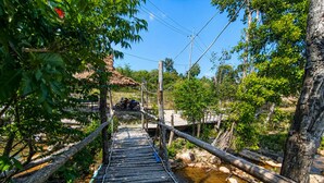 Eine rustikale Holzbrücke über einem Bach, im Hintergrund ein Gebäude mit einem Strohdach.