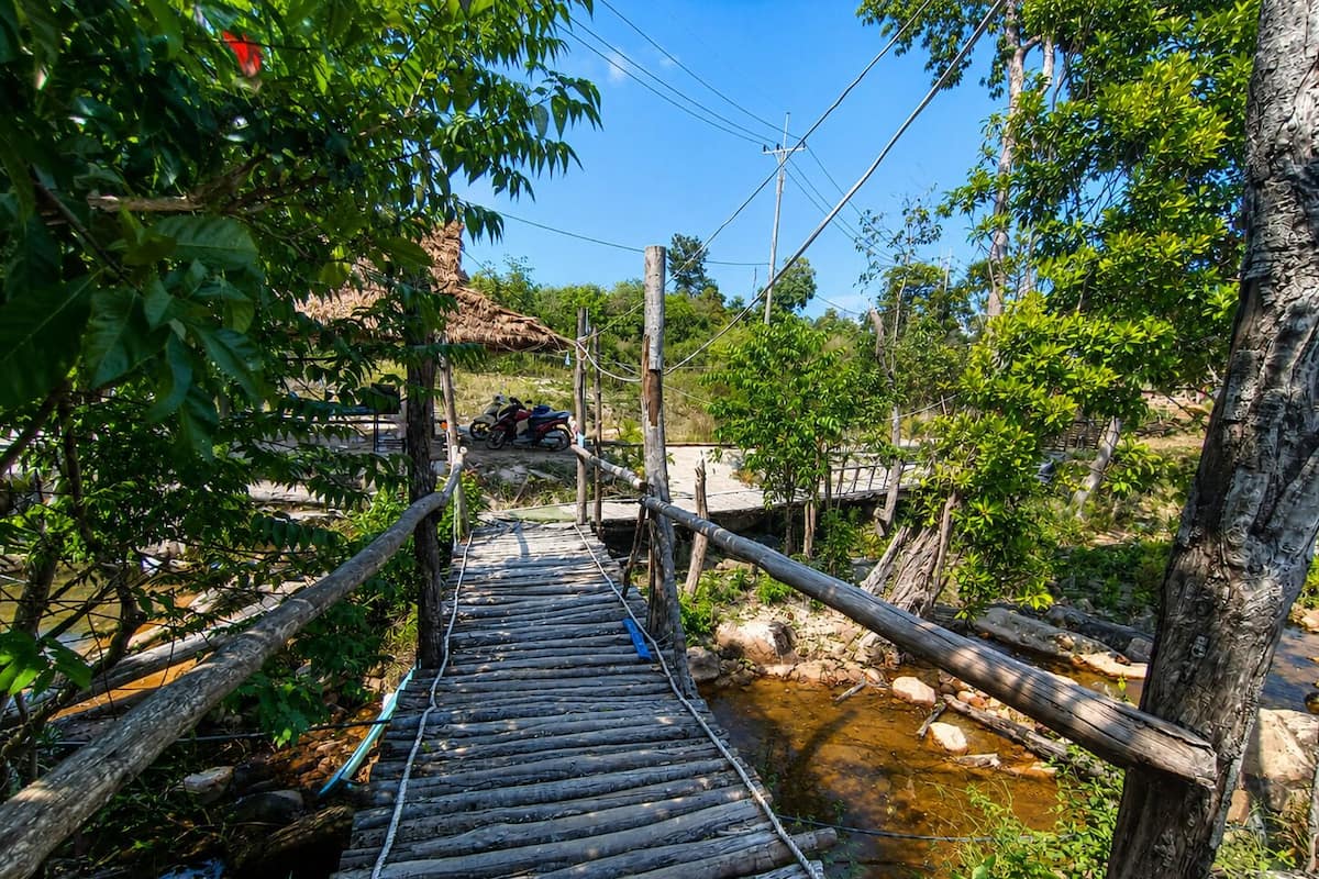 A rustic wooden bridge over a stream with a thatched-roof structure in the background.