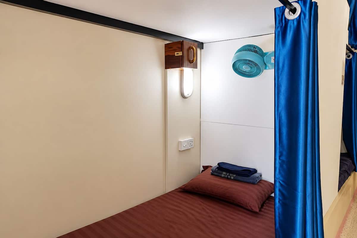 A small, single-bed room with a wooden headboard, a wall-mounted fan, and a power outlet.