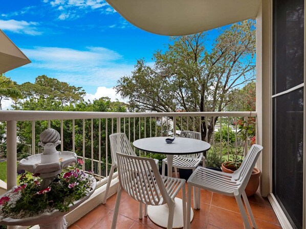 A balcony with a round table and four chairs, surrounded by a white railing, with a view of trees and a clear sky. - Relaxing Pool Hervey Bay Getaway Steps from Beach (Torquay)