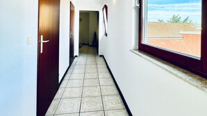 Un pasillo estrecho con suelo de baldosas, una puerta de madera a la izquierda y un ventanal grande a la derecha.
