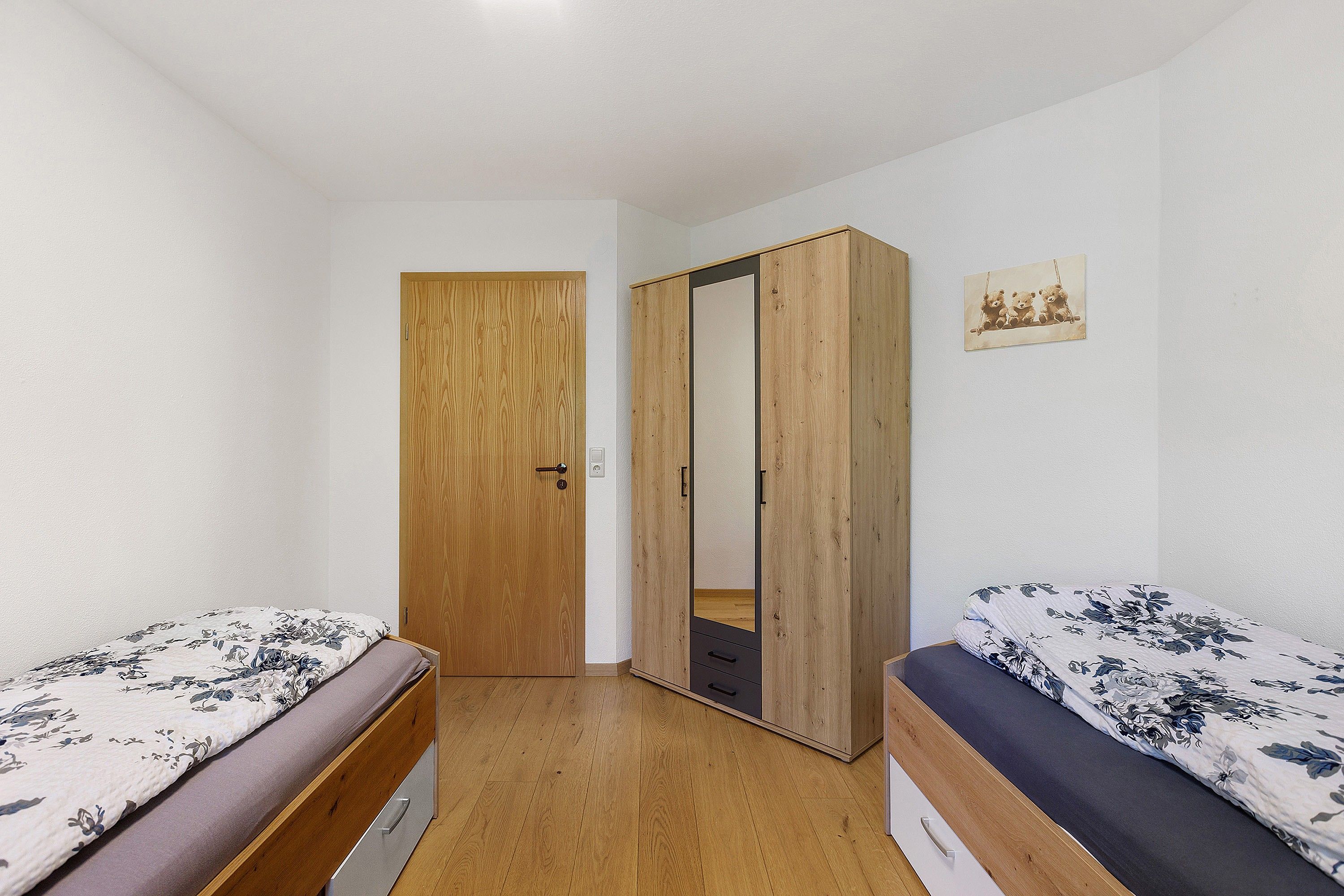 Une petite chambre avec un lit, un placard en bois, deux lits avec une literie à motifs floraux et un meuble avec miroir.
