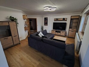 Una sala de estar moderna con un sofá oscuro, armarios de madera, un televisor de pantalla plana y un candelabro en el techo.