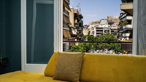 Un canapé jaune orné d’un coussin à motifs, dans une pièce dotée d’une grande fenêtre donnant sur un paysage urbain.