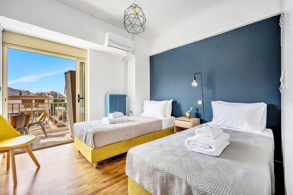 Une chambre d’hôtel avec deux lits, un balcon offrant une vue, une chaise jaune et un luminaire suspendu moderne.
