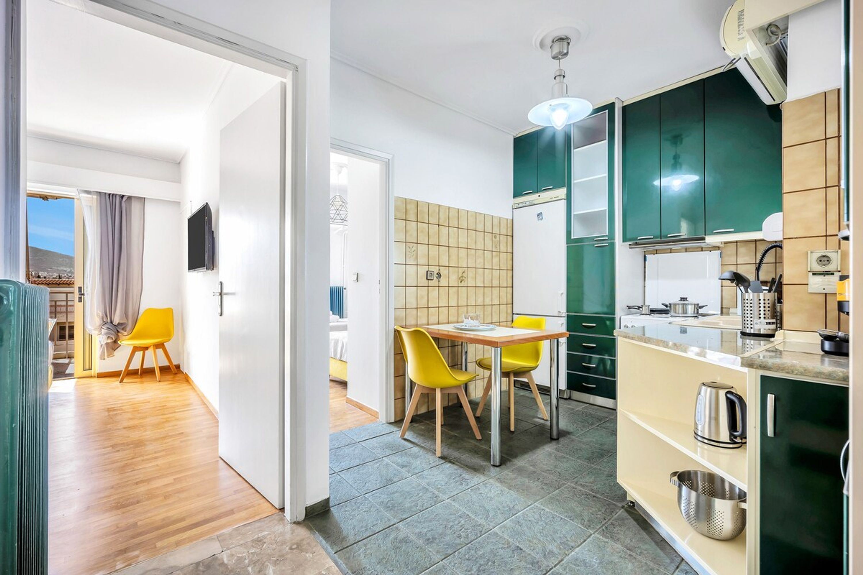 A modern kitchen with green cabinets, a dining area with yellow chairs, and a view of a balcony.