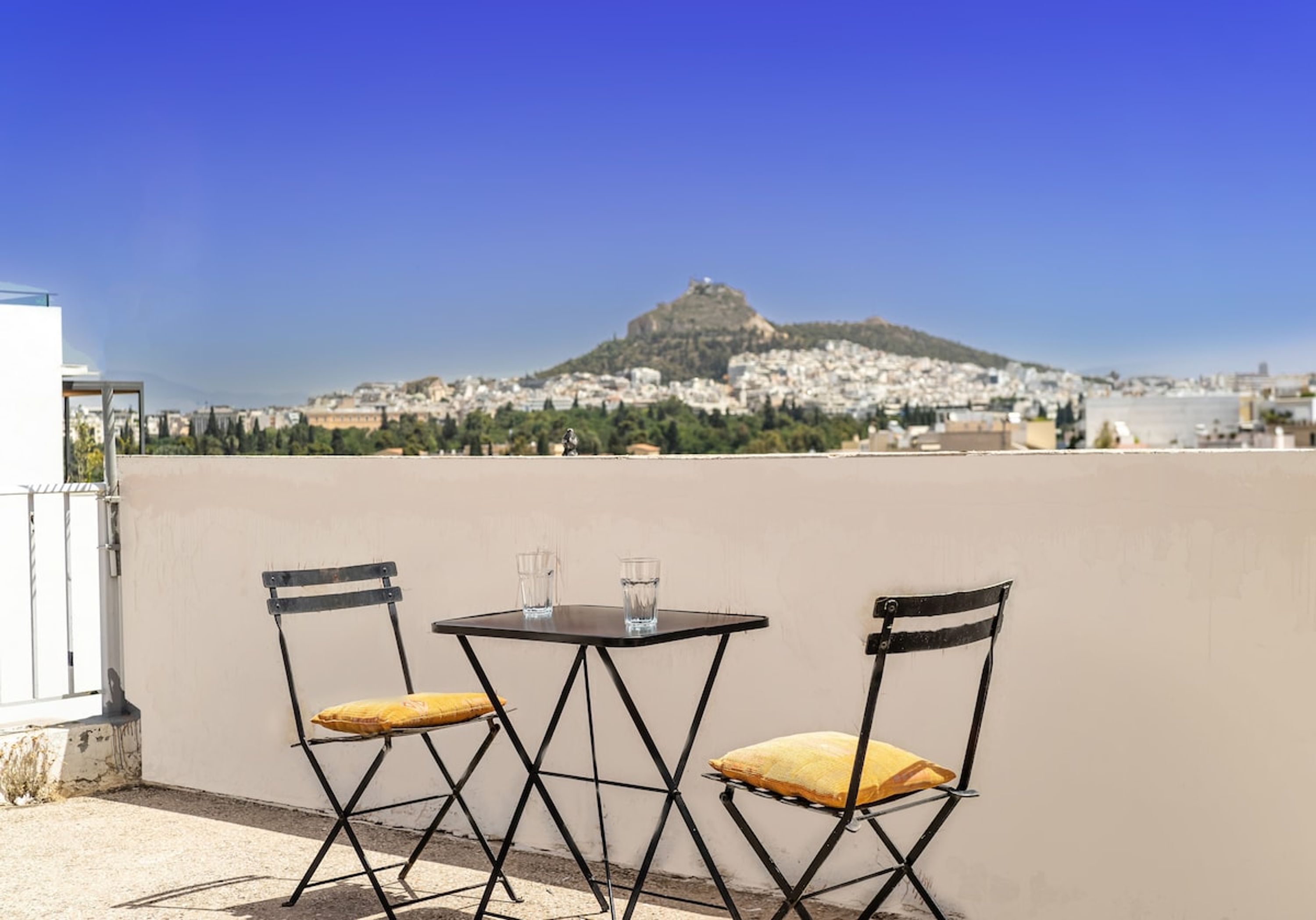 A rooftop terrace with two foldable chairs and a small table, overlooking a cityscape with a prominent hill in the background.