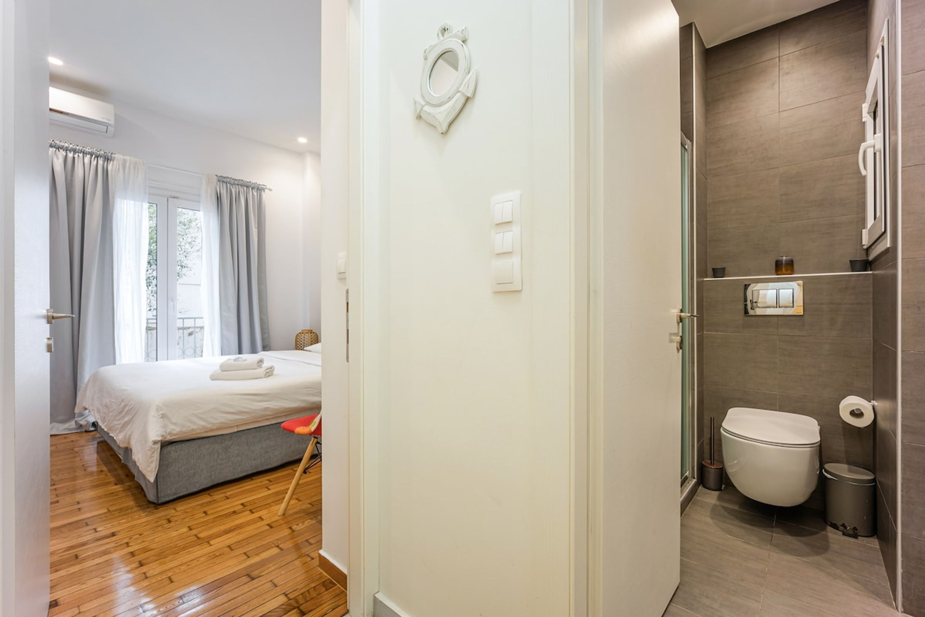 A hotel room with a wooden floor, a bed with grey bedding, a bedside table, and a window with curtains.