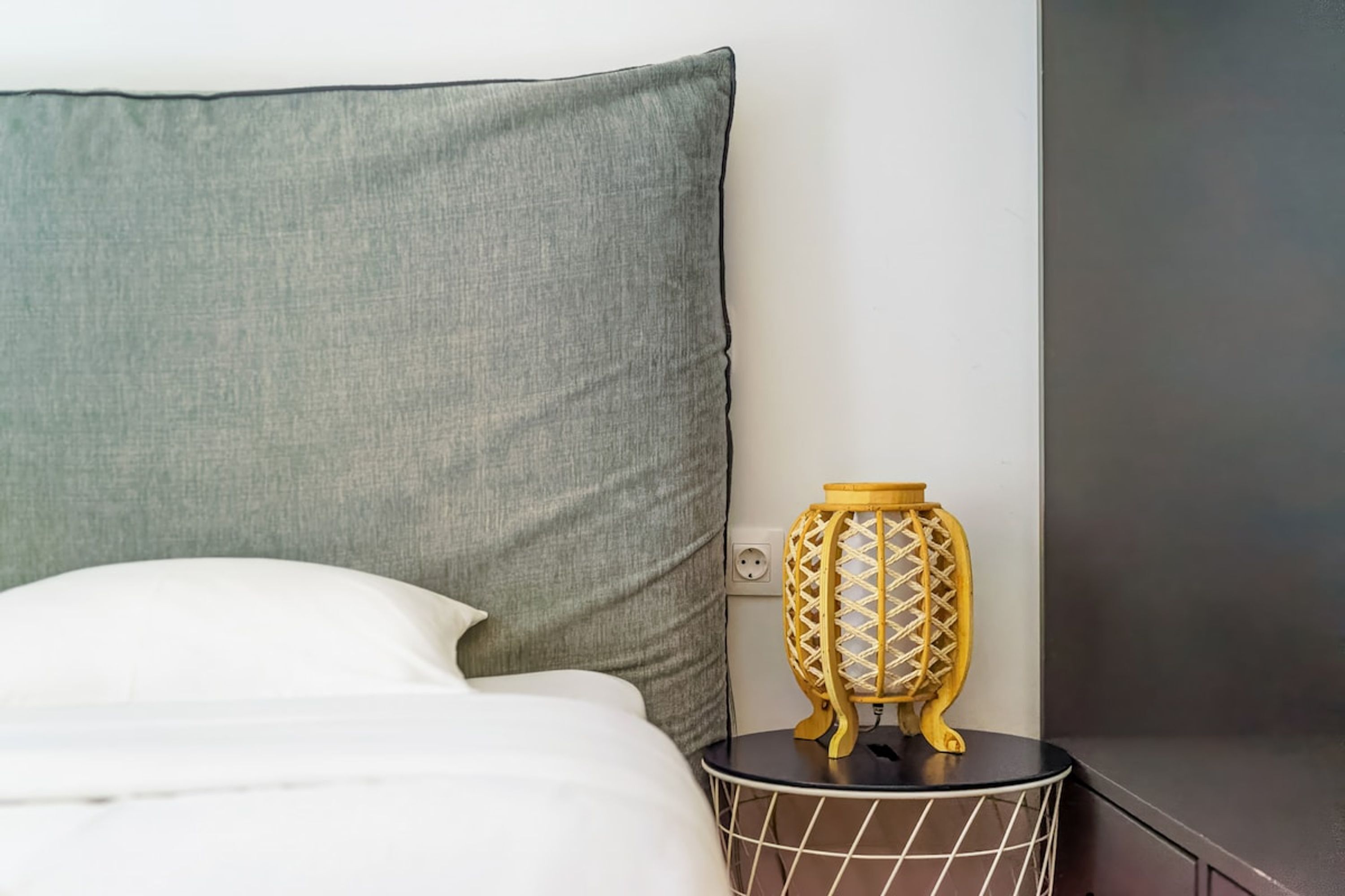 A bed with a grey headboard, a white pillow, and a small bedside table with a decorative lamp.