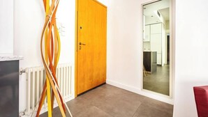 Un pasillo con puerta de madera, un radiador y un espejo que refleja una habitación con escritorio y silla.