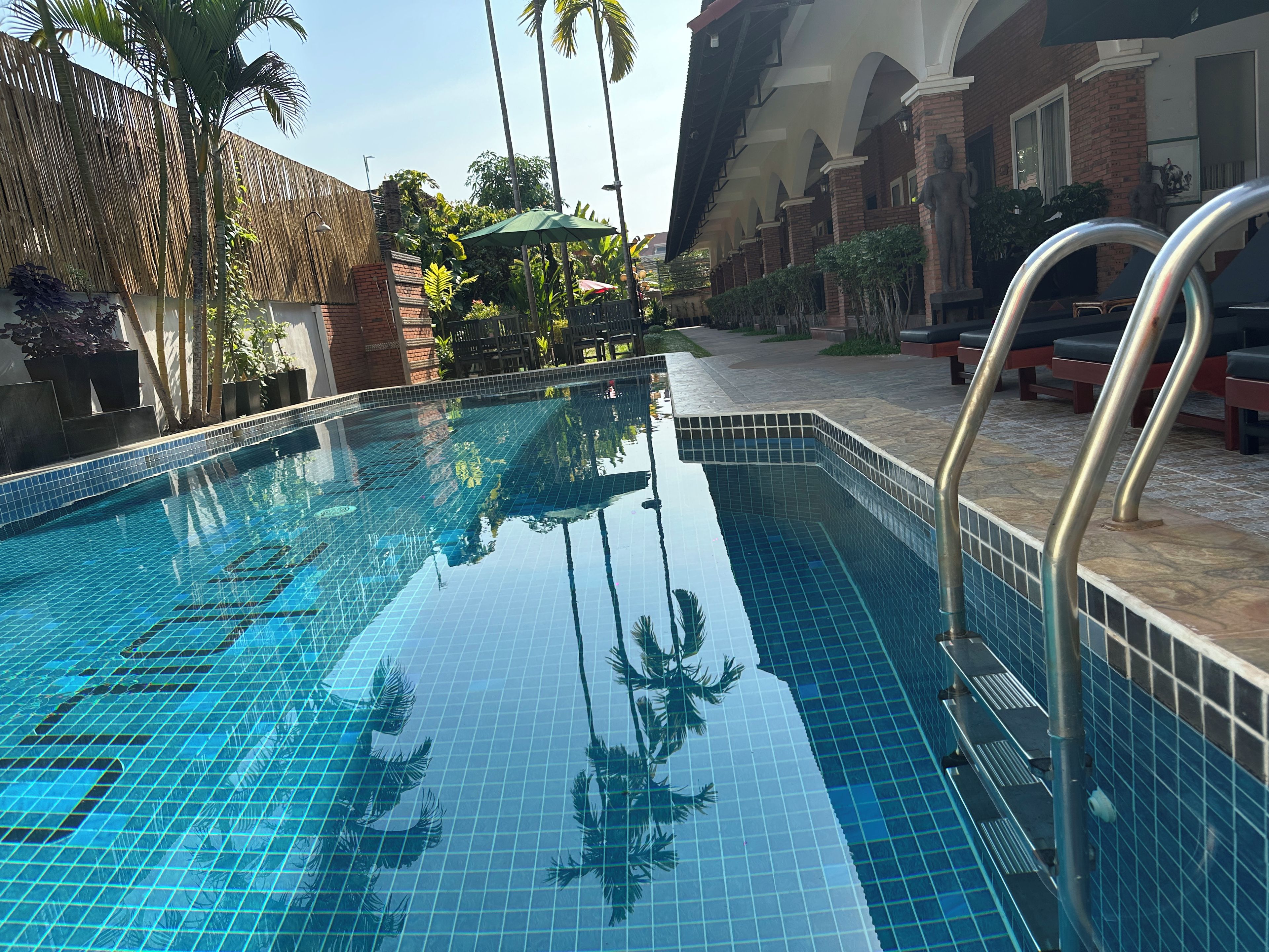 A swimming pool with clear blue water, surrounded by a brick building and palm trees.