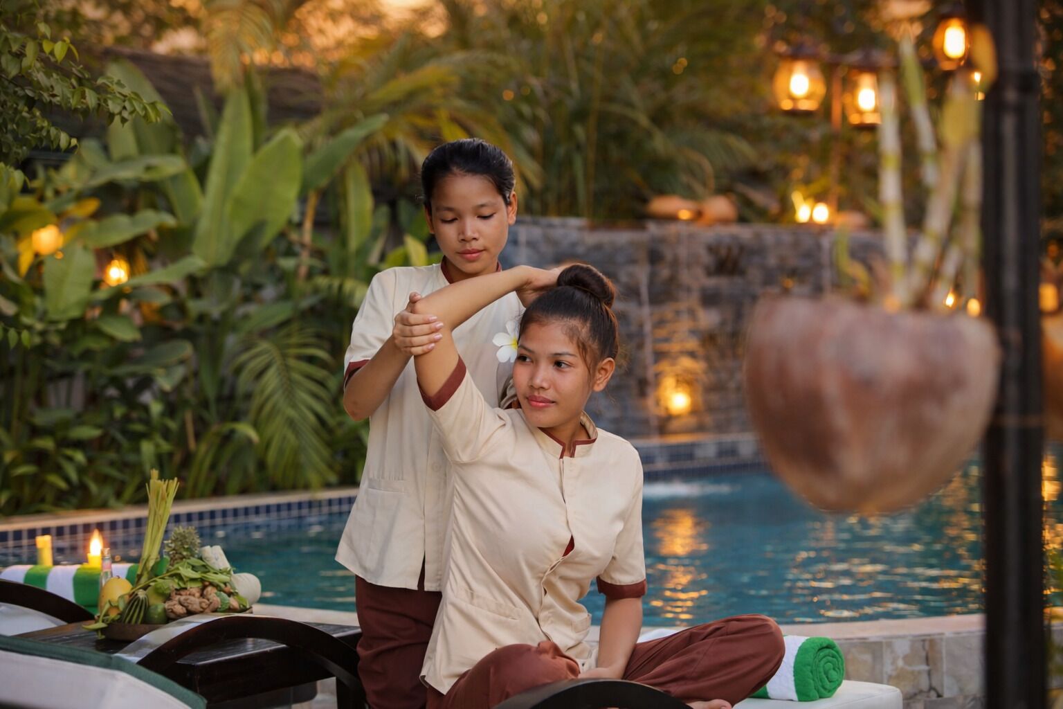 A person receiving a massage by another individual in a serene outdoor setting with a pool and lush greenery.