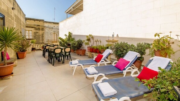 An outdoor patio with lounge chairs and tables, surrounded by potted plants and a white brick wall. - Ta Joseph (Xewkija)