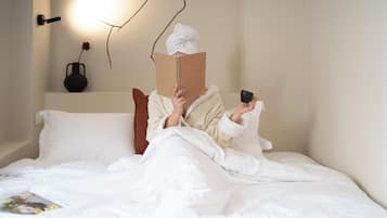 Uma cama com roupa de cama branca, um vaso preto com uma única flor e uma luminária fixada na parede.