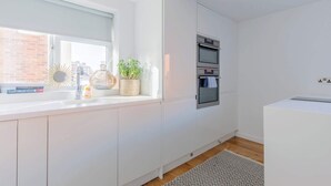 A modern kitchen with white cabinetry, a built-in oven, a window with a view of a brick building, and a small plant on the windowsill. - Chic 1BDR City Retreat with Views of the Shard (Greater London)
