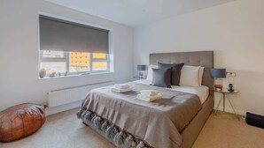A bedroom with a large window, a bed with pillows and a blanket, a side table with a lamp, and a radiator. - Chic 1BDR City Retreat with Views of the Shard (Greater London)