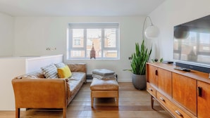 A modern living room with a sofa, ottoman, wooden cabinet, TV, and a large window with a view of buildings. - Chic 1BDR City Retreat with Views of the Shard (Greater London)