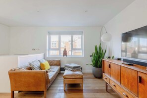 A modern living room with a sofa, ottoman, wooden cabinet, TV, and a large window with a view of buildings. - Chic 1BDR City Retreat with Views of the Shard (Greater London)