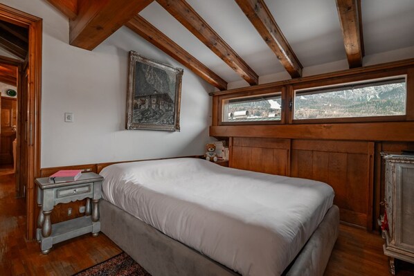 Quarto com cama, vigas de madeira, janela com vista, cómoda e um quadro na parede.
