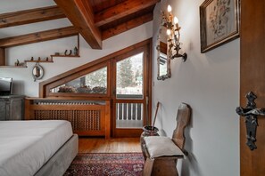 A bedroom with a sloped ceiling, wooden beams, a bed, a radiator, a window, a mirror, and a door. - GuestHost - Charming 45 sqm apartment, with fine wooden finishes and mountain view, ideal for two guests, located on the 4th floor of a characteristic building. The property is located in the heart of Cortina, along the famous Corso Italia, a 7-minute wa (Cortina d'Ampezzo)