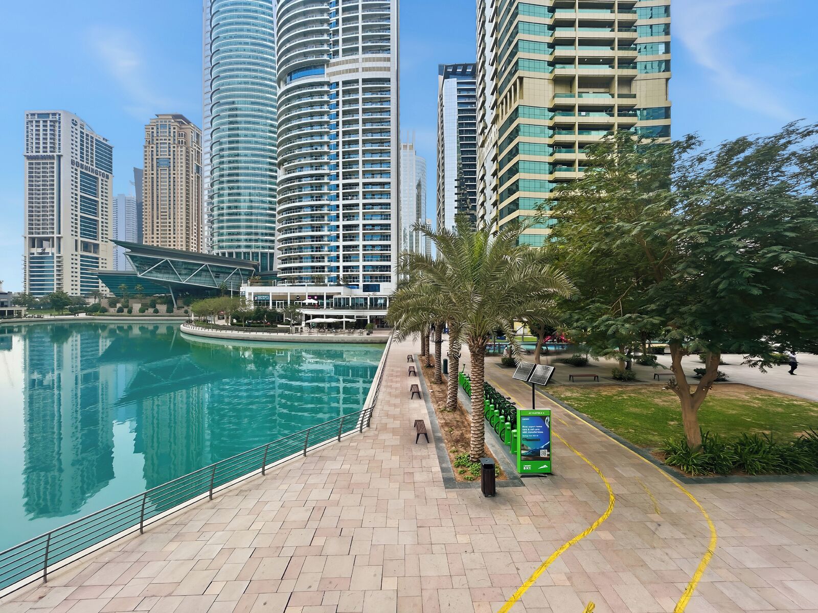 Eine Uferpromenade mit einem Spiegelteich, modernen Hochhäusern und einer Grünanlage.