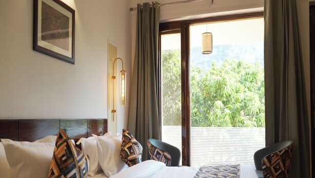 A hotel room with a large bed, two chairs, a wall-mounted light, a window with curtains, and a view of greenery outside. - The Whispering Oaks Corbett (Ramnagar)