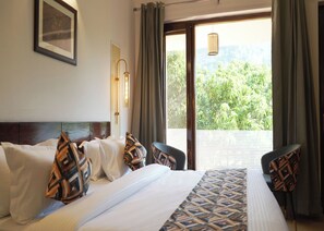 Camera d'albergo con un letto grande, due poltrone, una lampada a parete, una finestra con tende e vista sul verde.