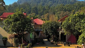 A courtyard with outdoor seating, surrounded by buildings with red roofs and lush greenery. - The Whispering Oaks Corbett (Ramnagar)