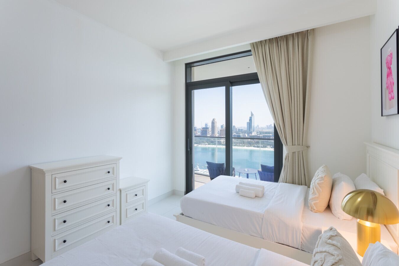 A hotel room with two beds, a white dresser, a large window with a city view, and a gold lamp.