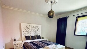 A bedroom with a large bed, a decorative headboard, a nightstand, a hanging light fixture, and a window with a view. - Riad Sahara Experience • Swimming pool (Souss-Massa Belfaa)