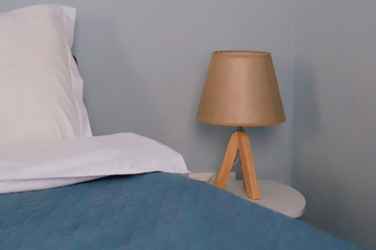 A bed with white bedding and a blue blanket, a wooden bedside table with a lamp, and a light blue wall.