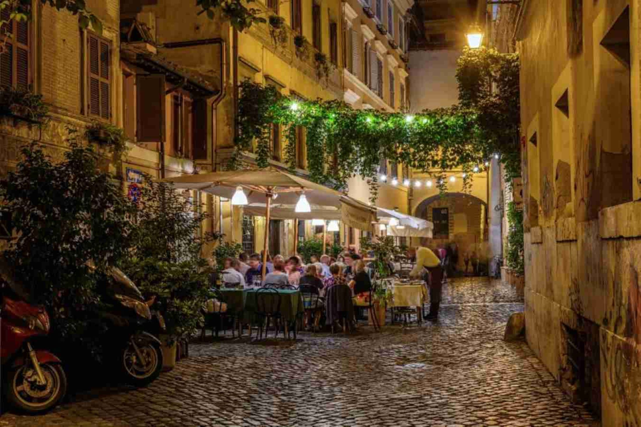 A cobblestone alley with outdoor seating, greenery, and a motorcycle.