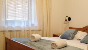 Een keurig opgemaakt bed met twee opgerolde handdoeken, een bedlamp en een raam met zonwering.