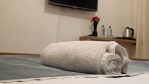 Een keurig opgemaakt bed met een opgerolde witte handdoek, een flatscreen televisie aan de muur, een klein houten nachtkastje met een vaas en flessen, en een waterkoker.