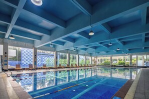 Une piscine intérieure avec des couloirs bien délimités, des chaises longues en bois et de grandes fenêtres donnant sur un paysage verdoyant.