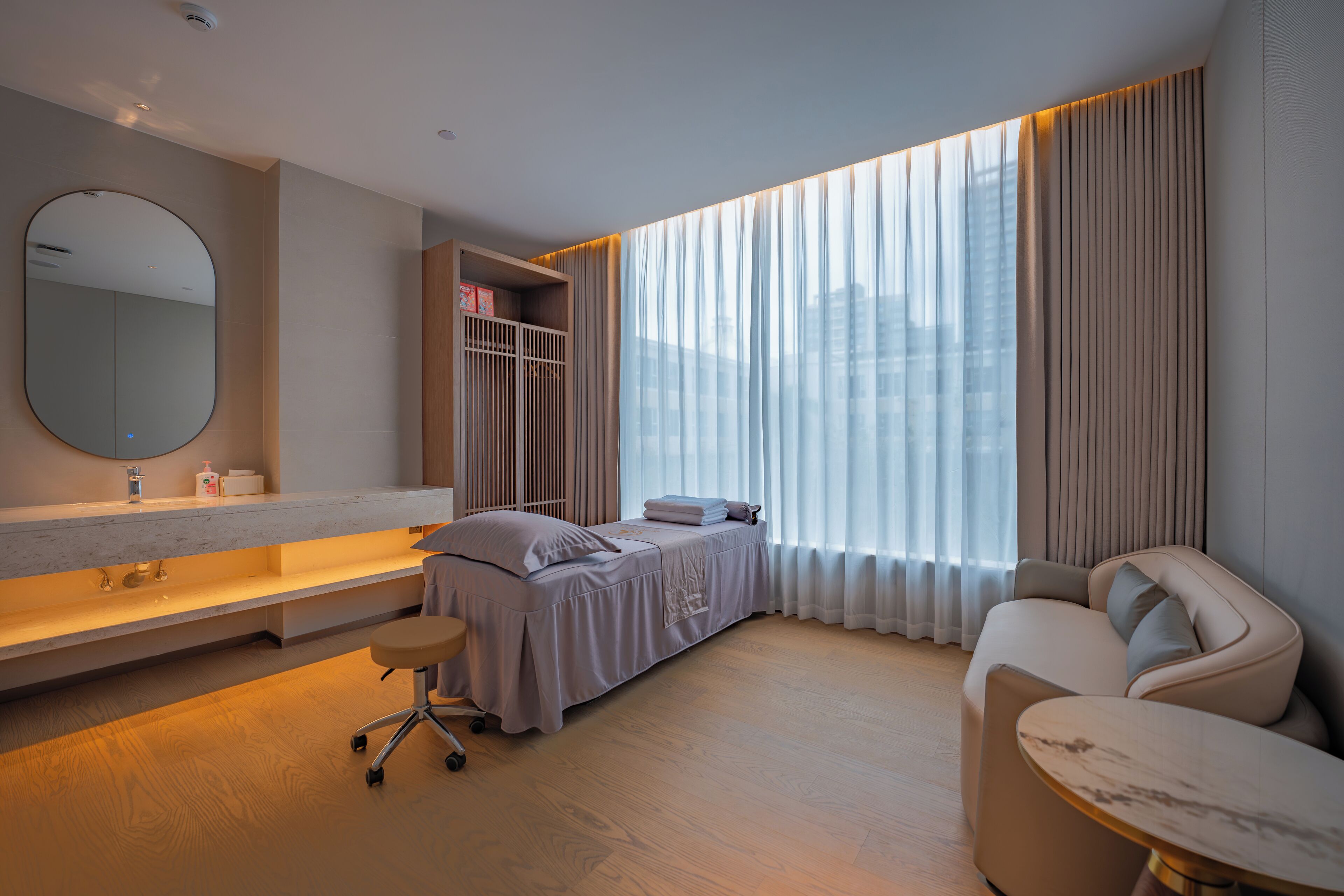 Una habitación de hotel moderna con una cama grande, una zona de estar, un tocador con espejo y un amplio ventanal con cortinas.