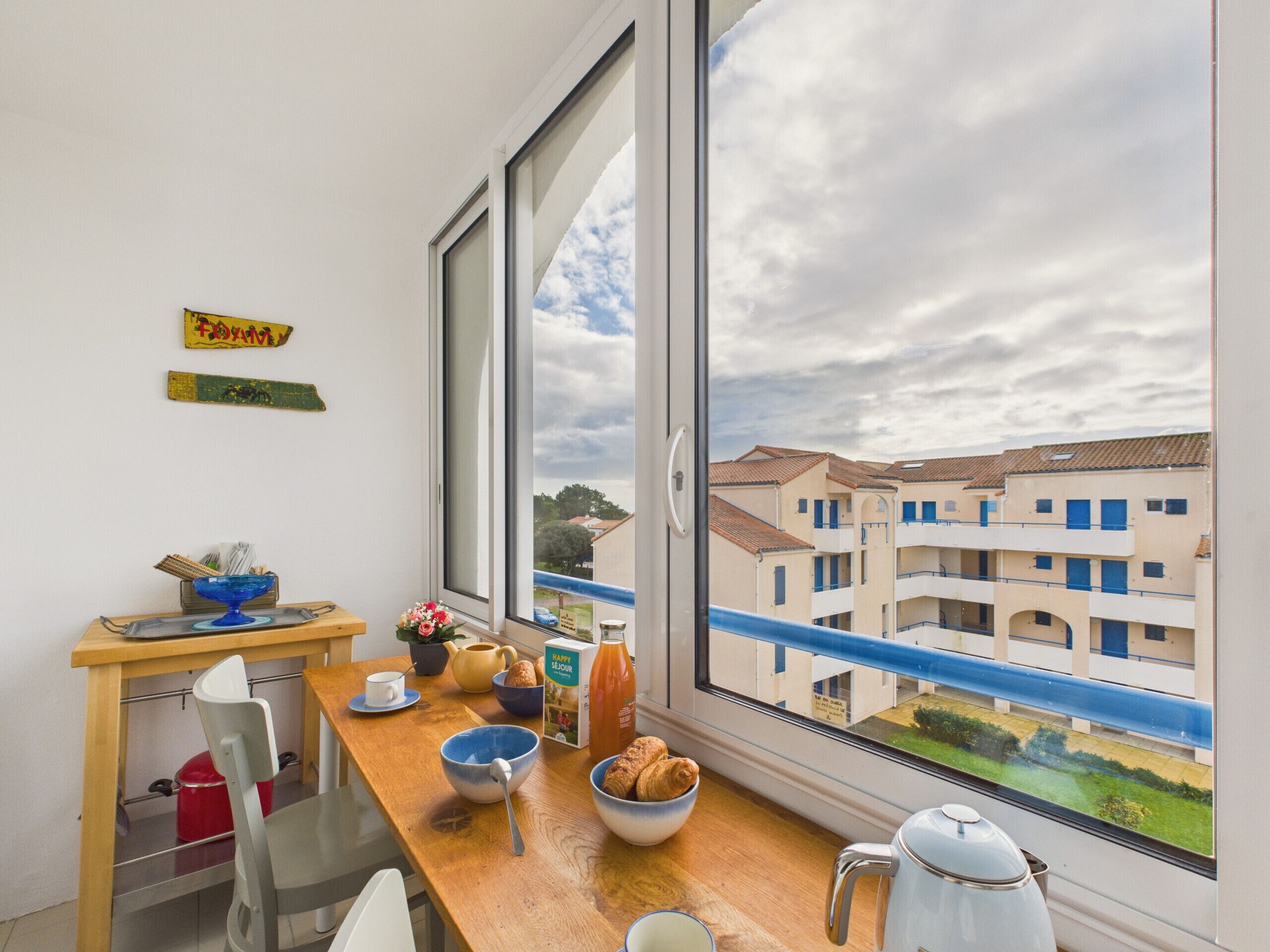 Ein Zimmer mit großem Fenster und Blick auf eine Wohngegend, einem Holztisch mit Frühstücksgebäck und einem Stuhl.