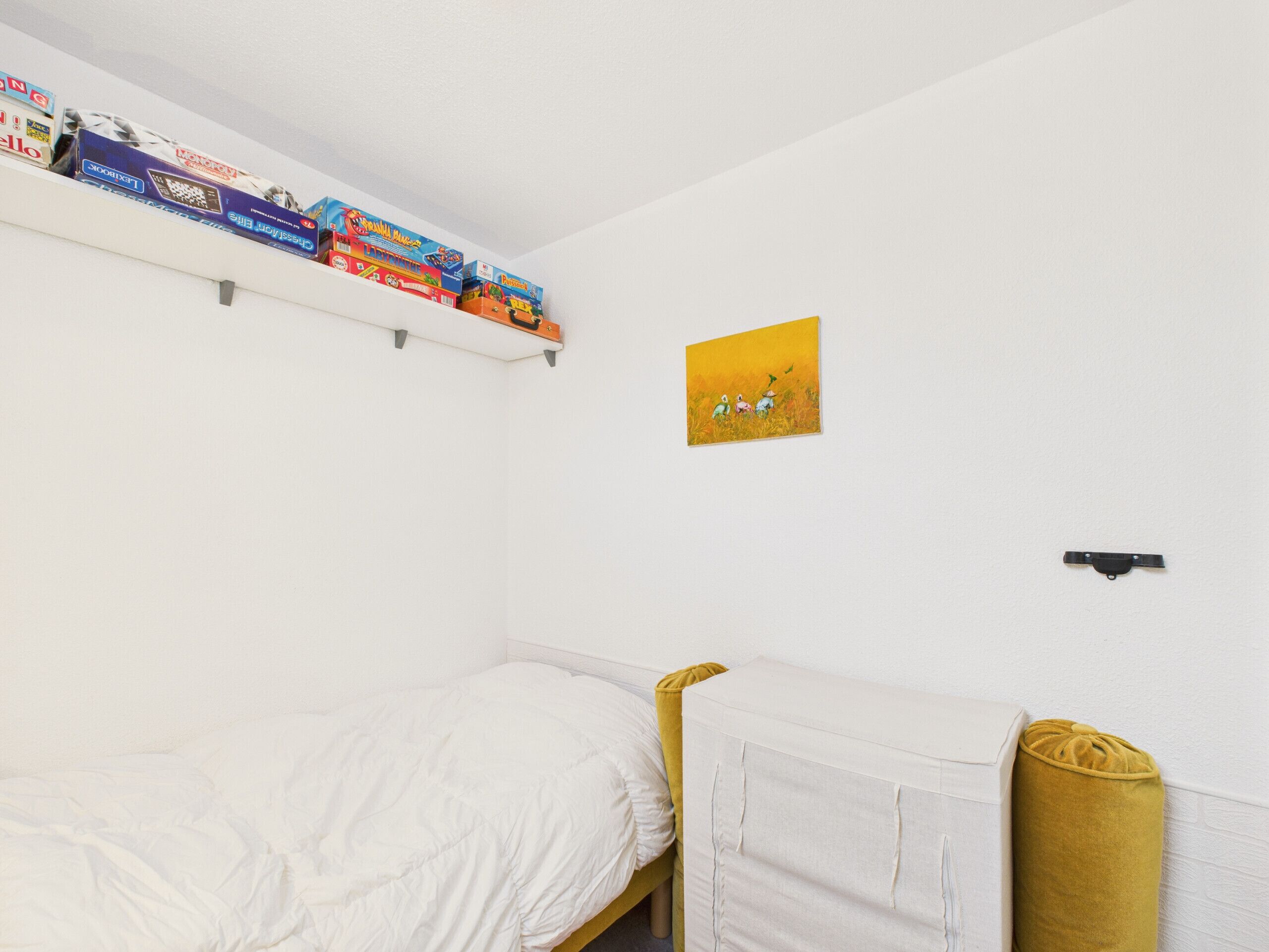 Une chambre à coucher avec un lit, une étagère remplie de livres, un tableau et un petit objet noir accroché au mur.
