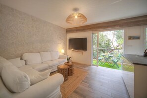 A modern living room with a white sofa, a wooden coffee table, a TV mounted on the wall, and a sliding glass door leading to an outdoor area with a table and chairs. - 2 Bedrooms Balcony garden view-Carnot A2B266 (Cannes)