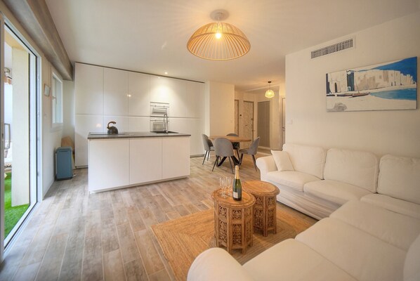 Un saló modern amb un sofà blanc, una zona de menjador amb taula i cadires, una cuina amb armaris blancs i una finestra gran.