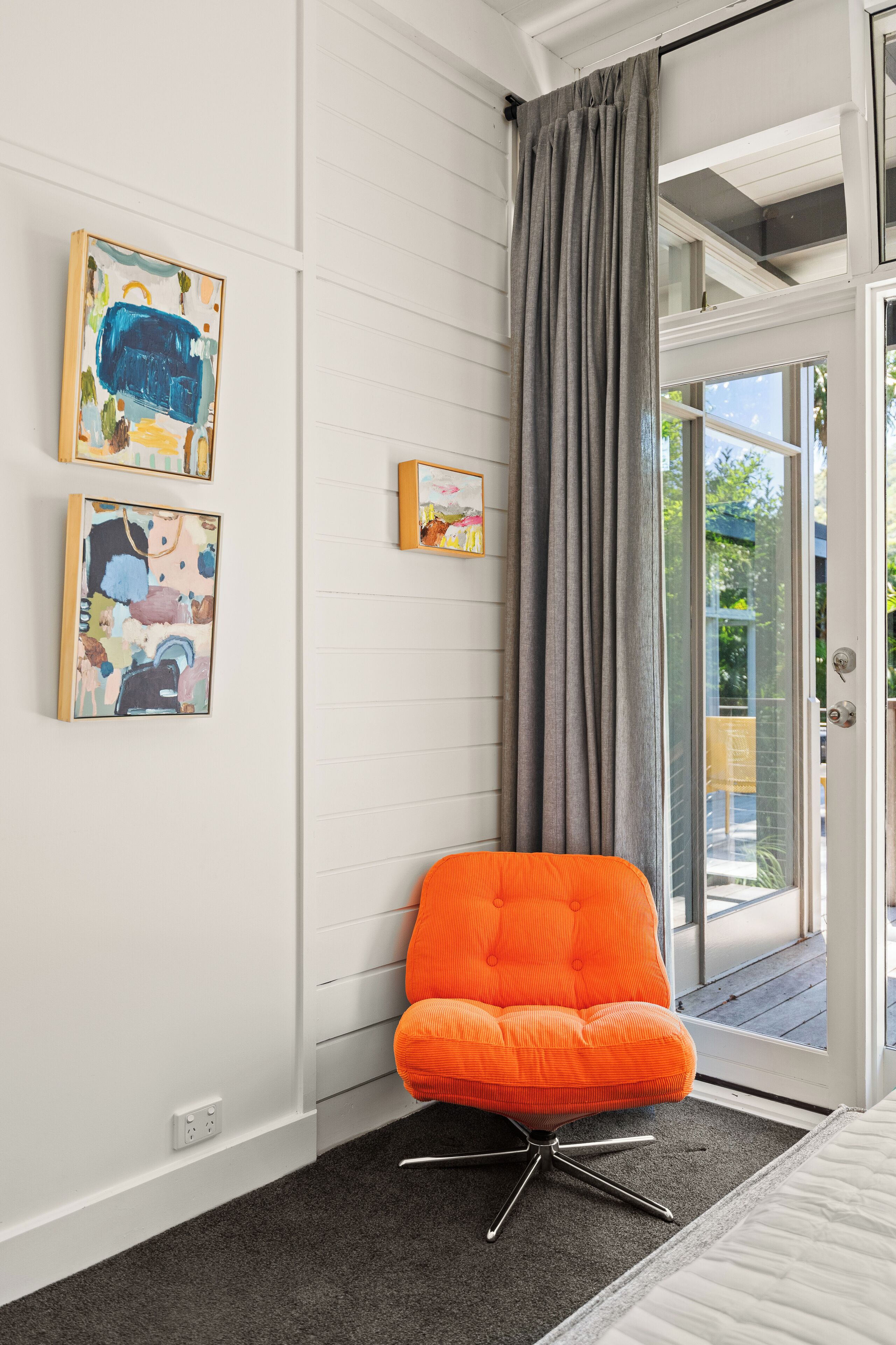 A room with a white wall, a gray curtain, and a bright orange chair. There are three framed artworks on the wall.