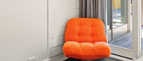 A room with a white wall, a gray curtain, and a bright orange chair. There are three framed artworks on the wall.