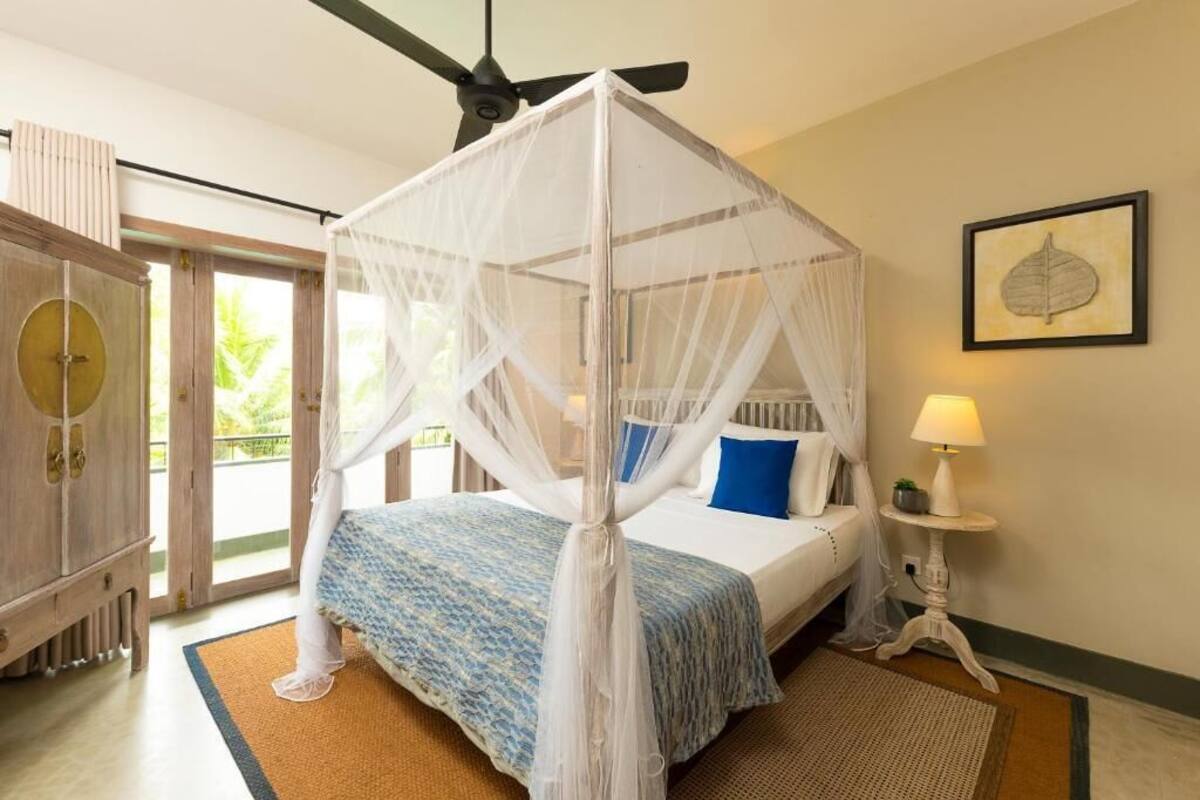 Un dormitorio con cama con dosel, ventilador de techo, lámpara de noche y vista al exterior a través de un ventanal.