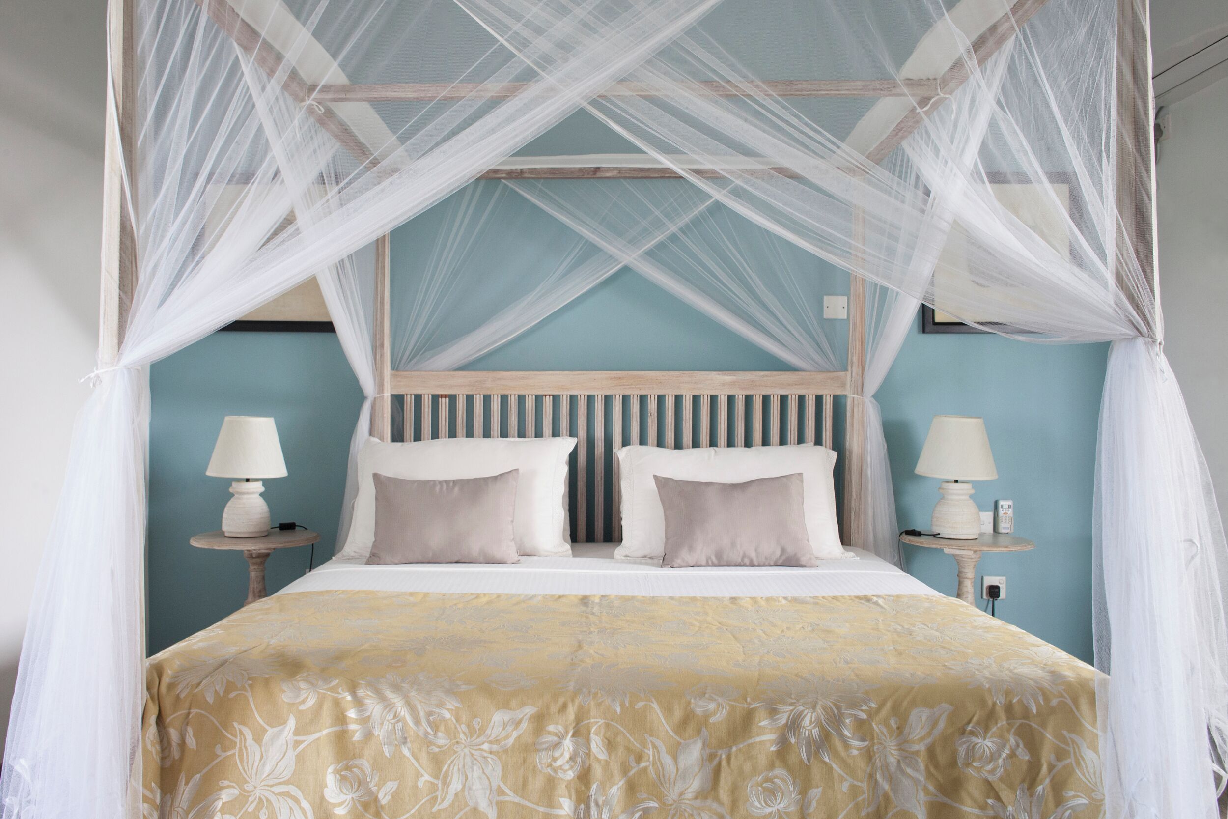 Un lit à baldaquin à quatre poteaux, surmonté d’une couvre-lit à motifs floraux, deux tables de chevet avec des lampes et une petite table fixée au mur, servant de table de nuit.