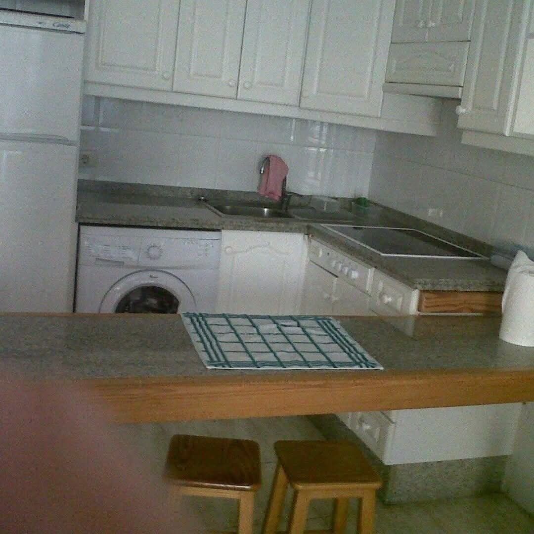 Een kleine keuken met een wasmachine, een spoelbak en een fornuis. Twee krukken en een tafeltje met een placemat met ruitmotief.