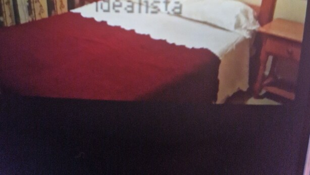 Una cama con cabecera de madera, una colcha roja y una mesita de noche con lámpara.