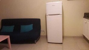 Un espacio habitable reducido con un sofá de color oscuro junto a una pared, un refrigerador blanco y una mesita.